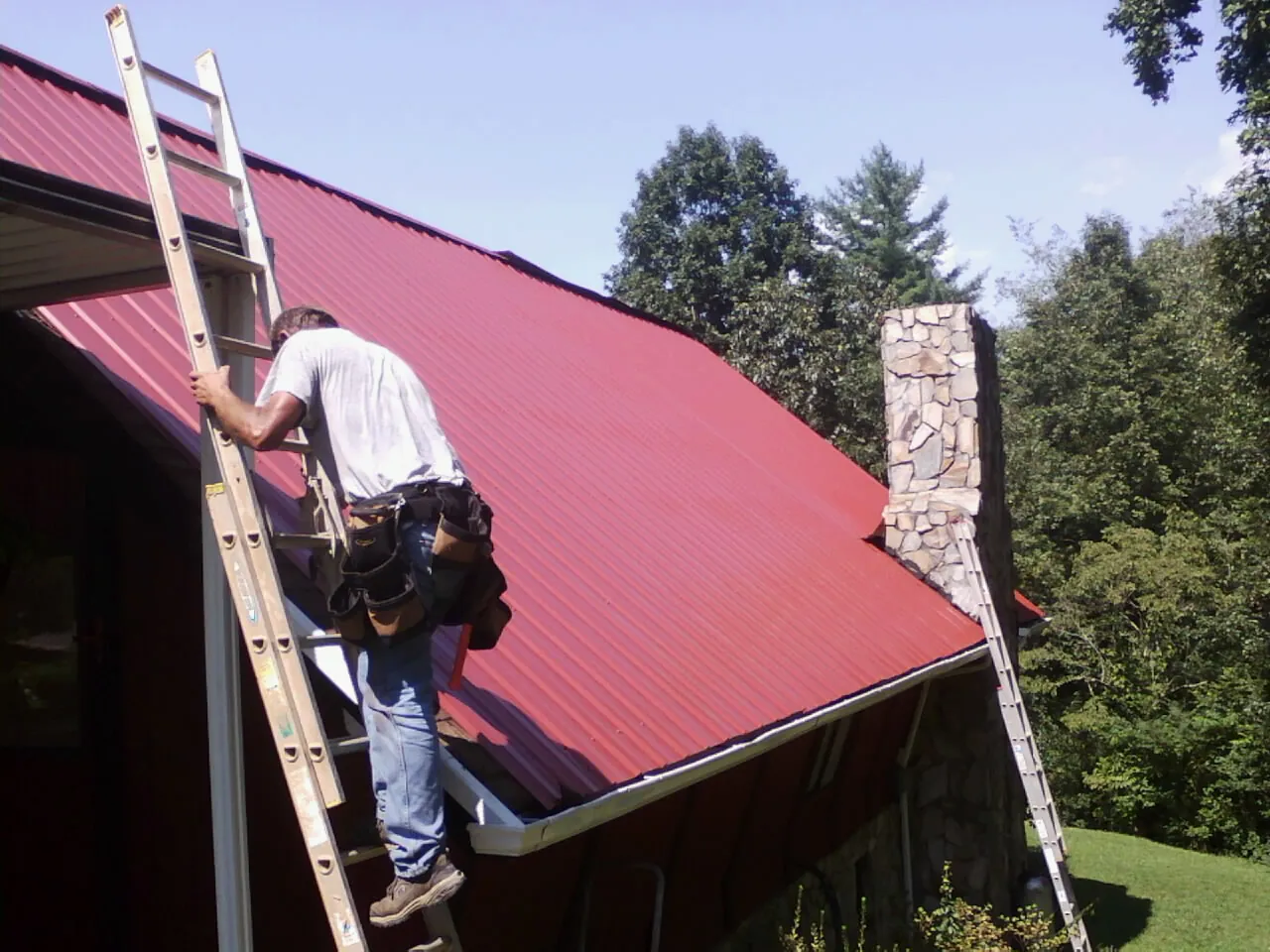Roofer on a ladder beside a red metal roof and stone chimney — Soffit & Fascia Repair by Roofing Brothers in Sutherland Springs
